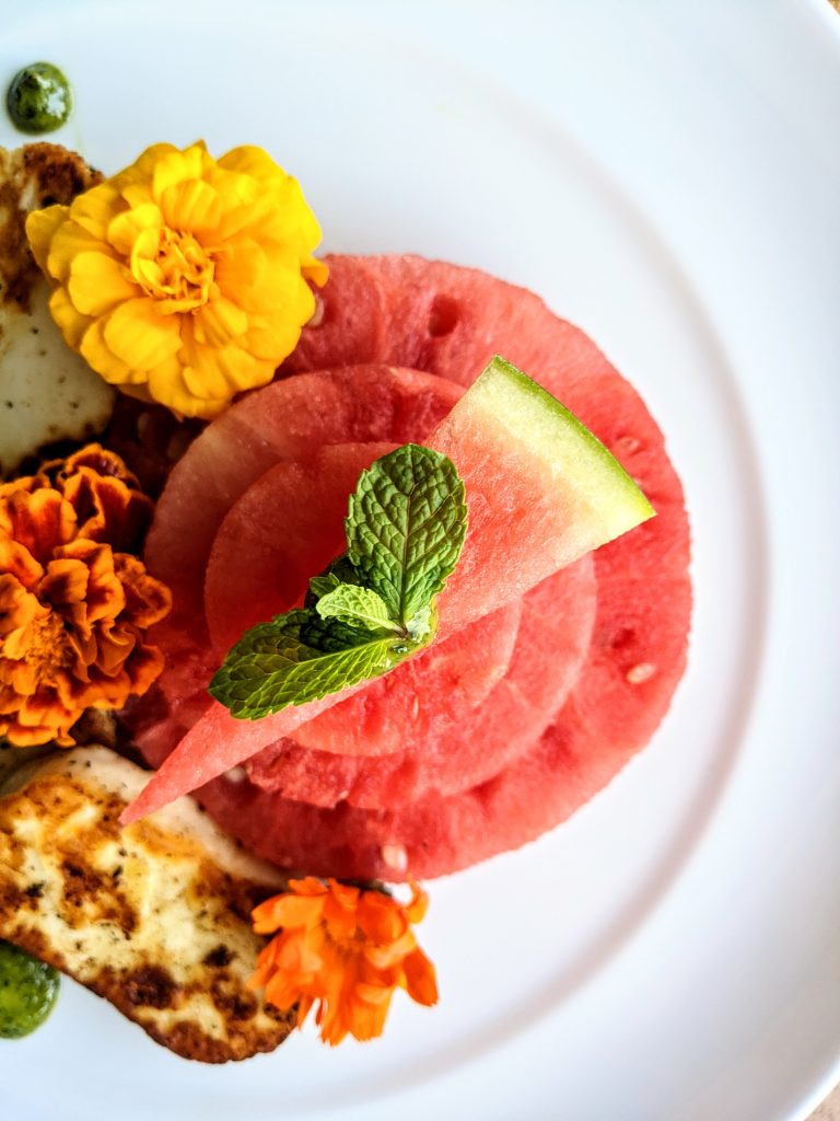 A close-up of circular watermelon, halloumi, edible flowers. and mint & cilantro pesto.