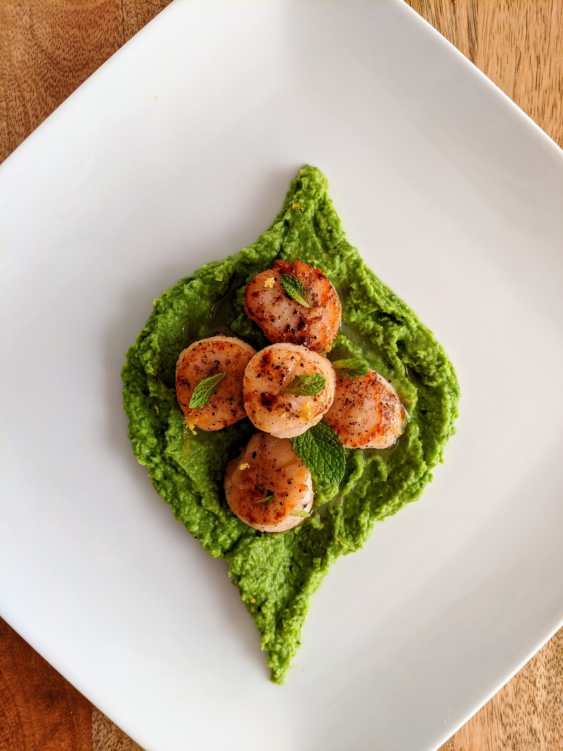 An aerial photo of five seared scallops on top of mint and pea purée in the shape of a mint leaf; garnished with fresh mint leaves.