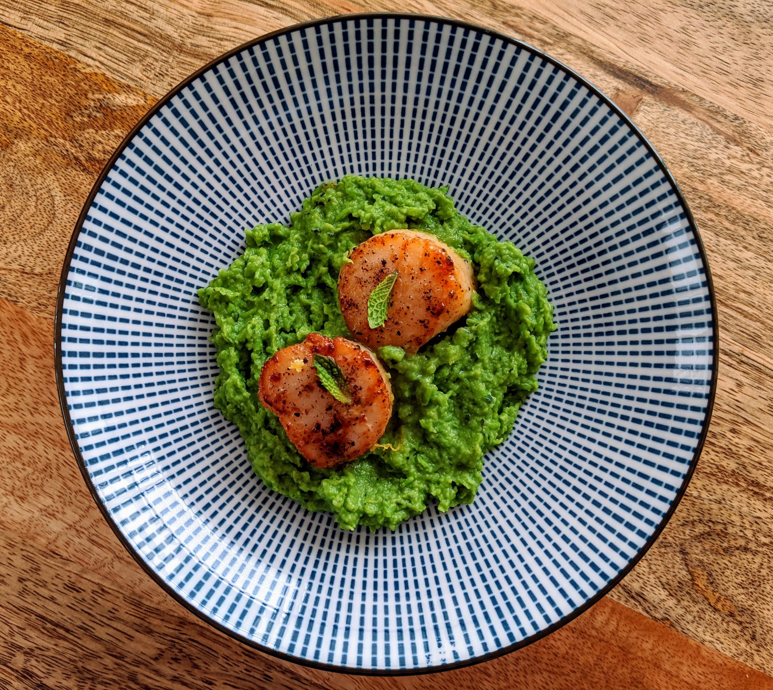 Two seared scallops on top of a circular bed of mint and pea purée; garnished with fresh mint leaves. This appetizer sized portion is served on a blue and white geometric print side plate.