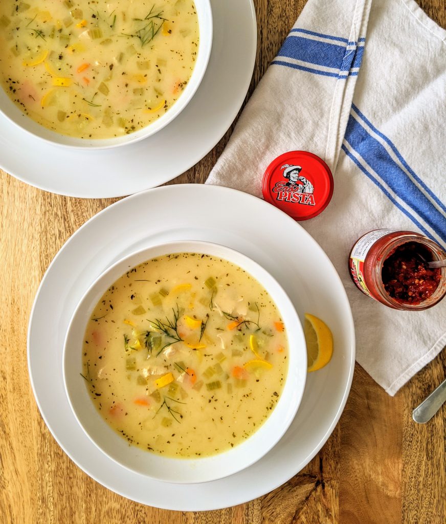 An aerial shot of two bowls of yellow colored chicken Mediterranean-style soup, garnished with fresh fill and lemon wedges. Served with Eros Pista hot chili to stir into the soup.