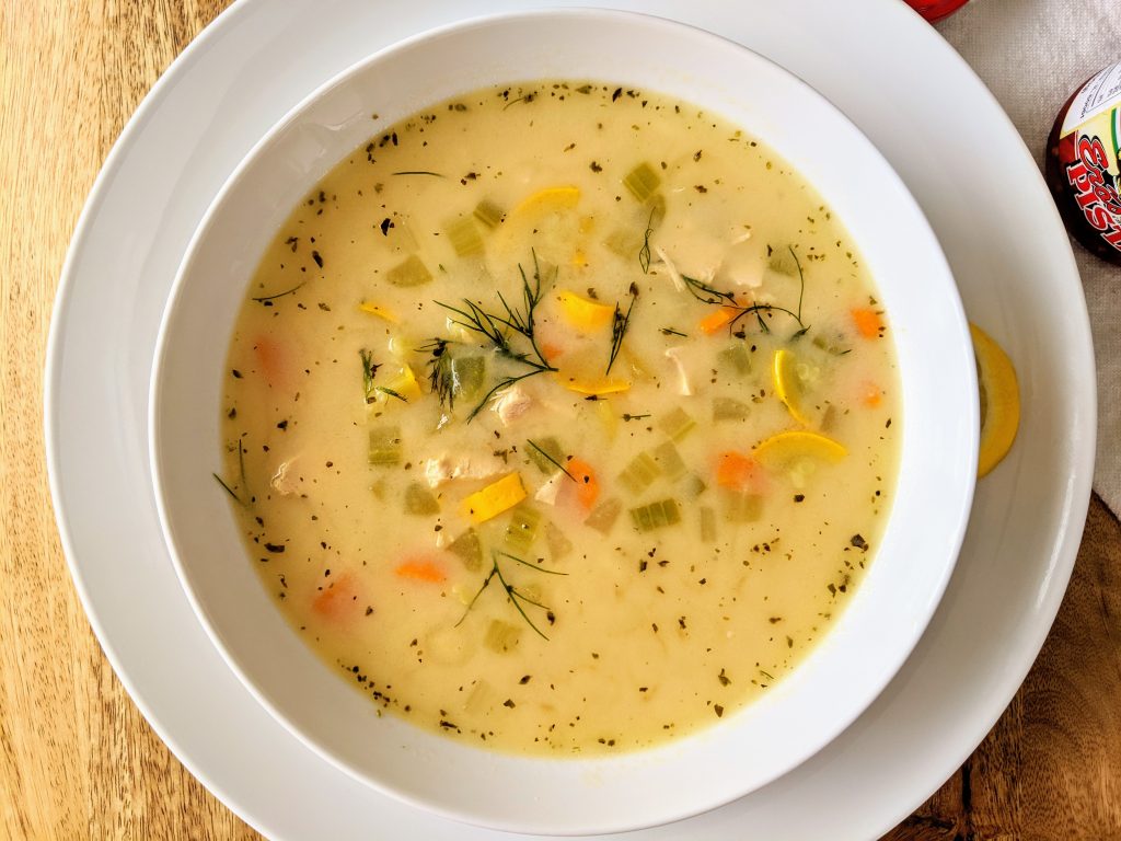 An aerial closeup of a Mediterranean chicken and vegetable soup; garnished with fresh dill and a lemon wedge.l
