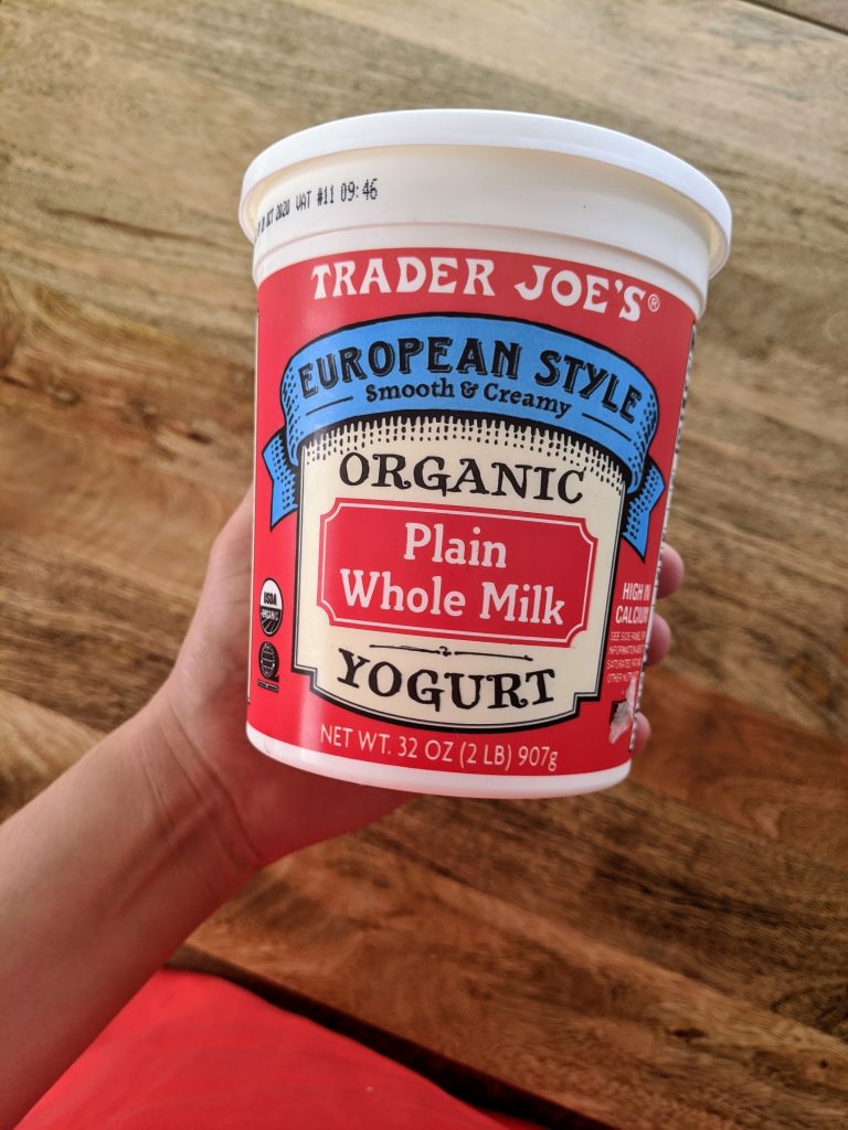 Me holding up a 32 oz container of Trader Joe's Organic European Style Plain Whole Milk Yogurt. The container has a red background and a blue banner style label.
