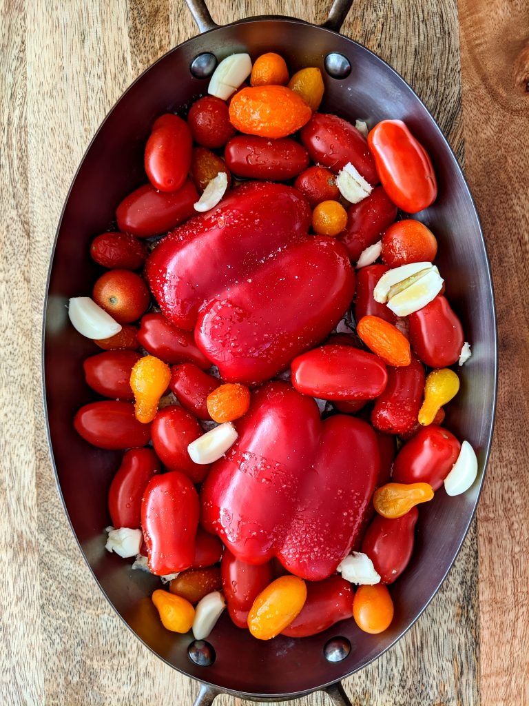 An oval baking dish with assorted small variety tomatoes, red pepper, and smashed garlic cloves, all ready to roast in the oven.