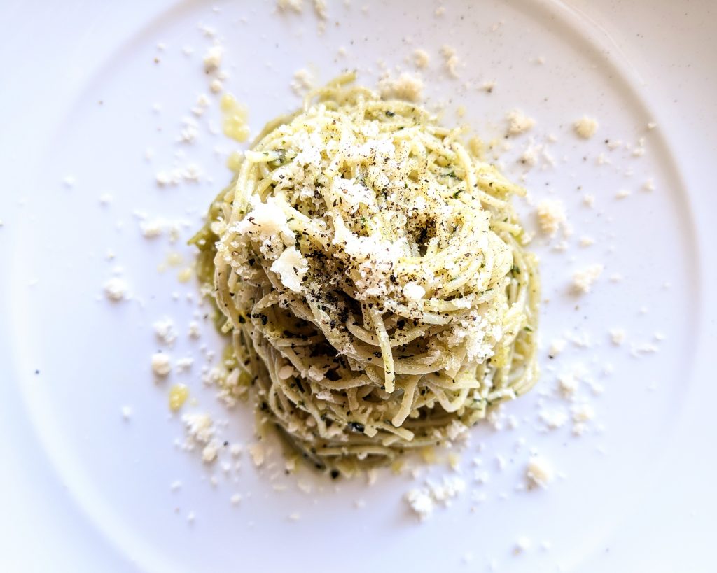 A closeup of a nest of pesto pasta, topped with finely grated Parmigiano-Reggiano and freshly ground black pepper.