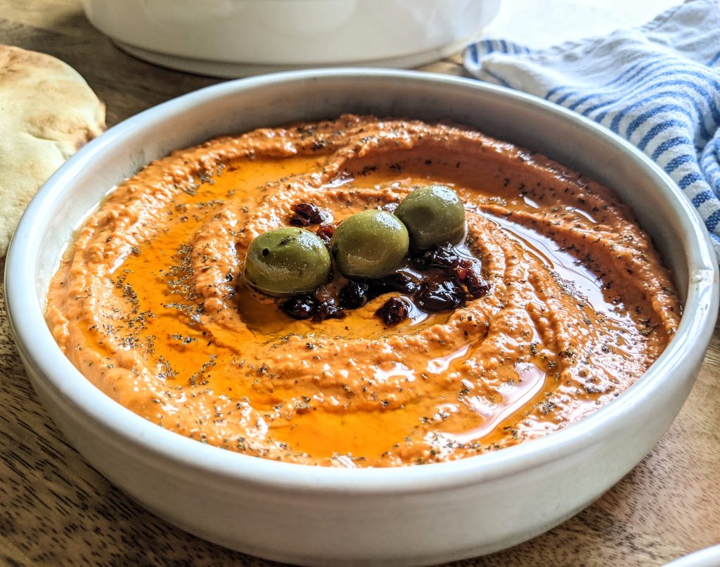 A closeup of roasted red pepper and sun-dried tomato hummus, garnished with green olives and chopped sun-dried tomatoes.