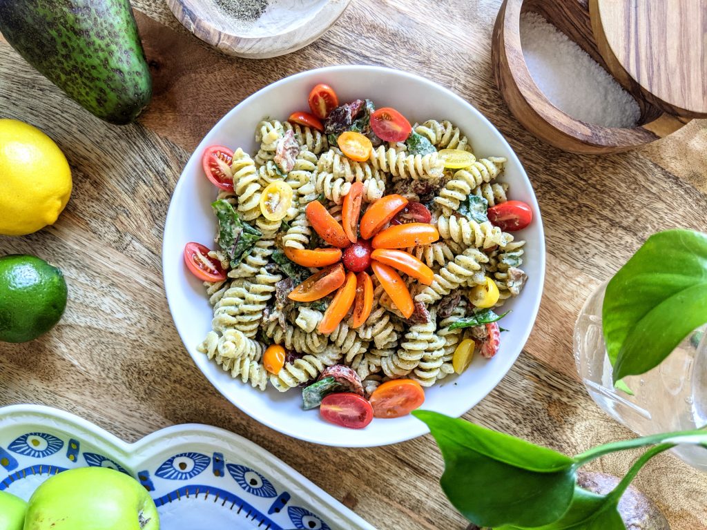 An aerial view of a bowl of BLT Pasta salad with rotini, crispy bacon, multi-colored tomatoes, and green baby Swiss Chard. There's a flower made out of orange and red tomatoes in the middle of the bowl.