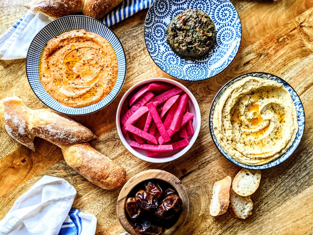 An aerial view of a table full of mezes. Middle Eastern pickled turnips being front and center, flanked by hummus, tirokafteri, olive tapenade, dates, and fresh bread.