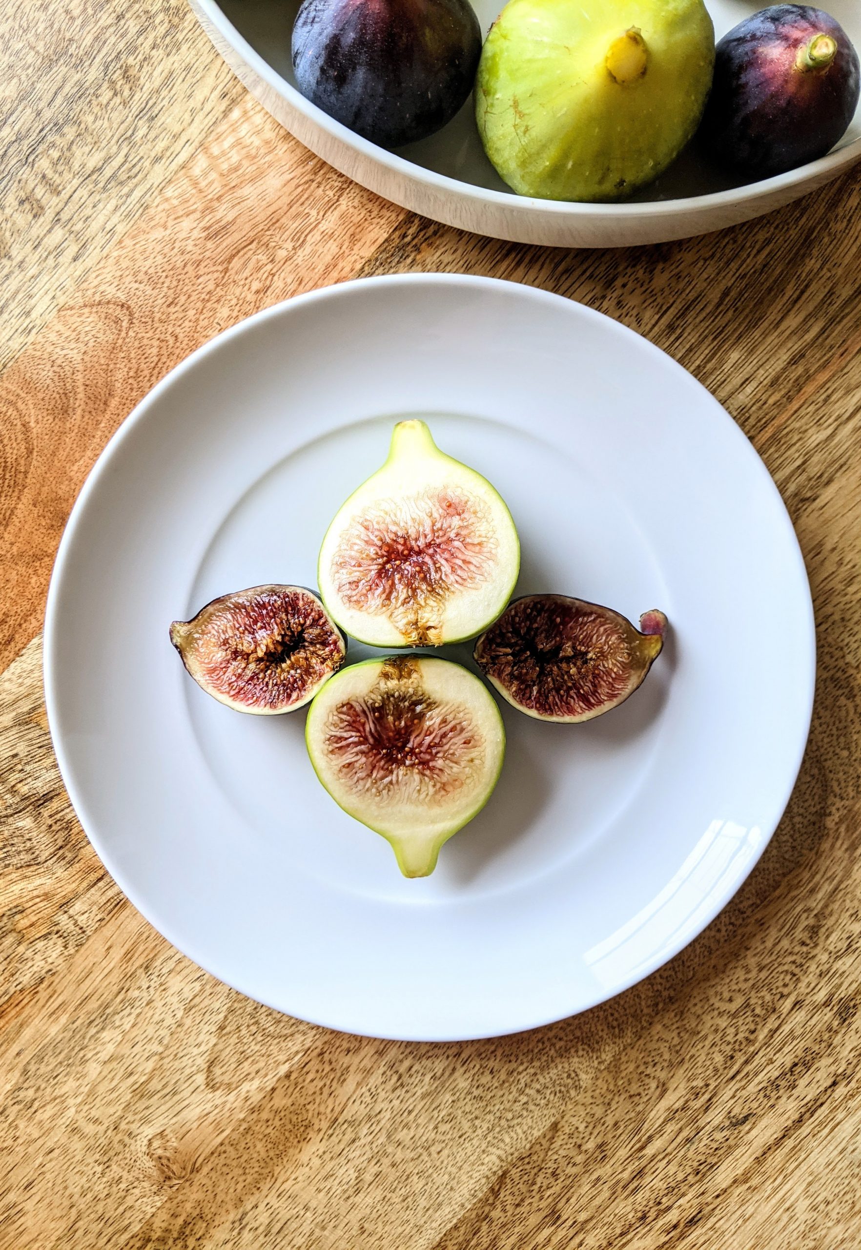 A fresh Tiger fig and a Mission fig sliced in half and placed flesh side up on a small white plate.