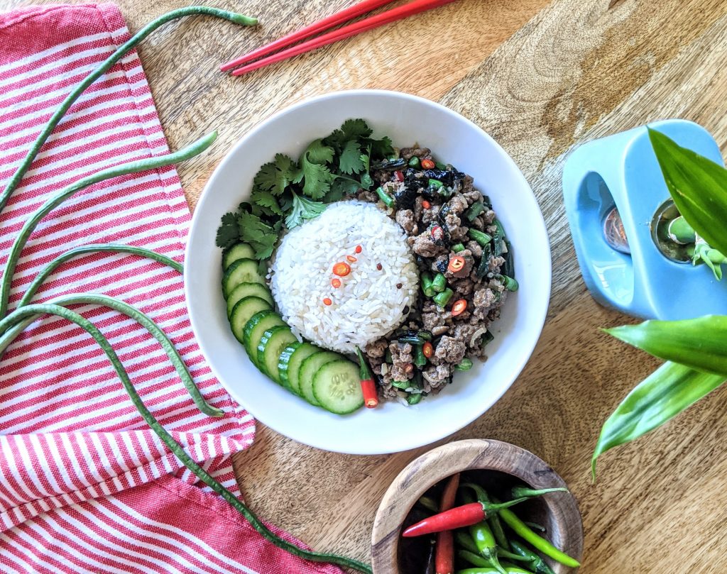 An aerial photo of Spicy Thai Basil Chicken, served with steamed Jasmine rice, sliced cucumbers, fresh cilantro, and fresh chilies. Long beans are visible on a red and white kitchen towel, a bamboo plant in a turquoise vase peeks into the frame, and a bowl of chilies sits next to the meal.
