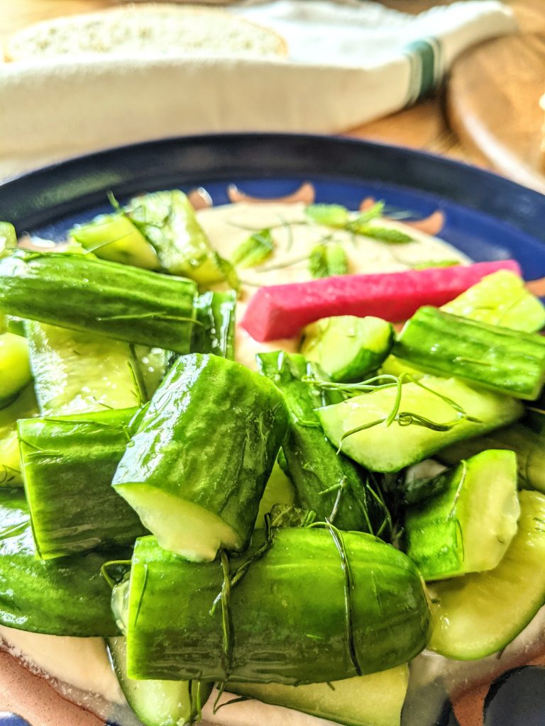 A close up shot of Smashed Cucumber Salad with Whipped Honey Feta, garnished with fresh dill & mint, and homemade pickled turnips.