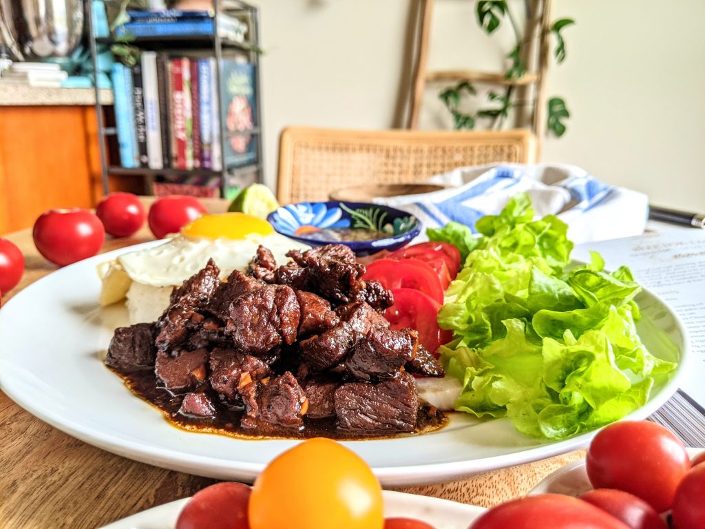 Cubed Beef Lok Lak served on a bed of green lettuce, with sliced tomato, Jasmine rice, a lightly fried egg with runny yolk, and lime pepper dipping sauce. 