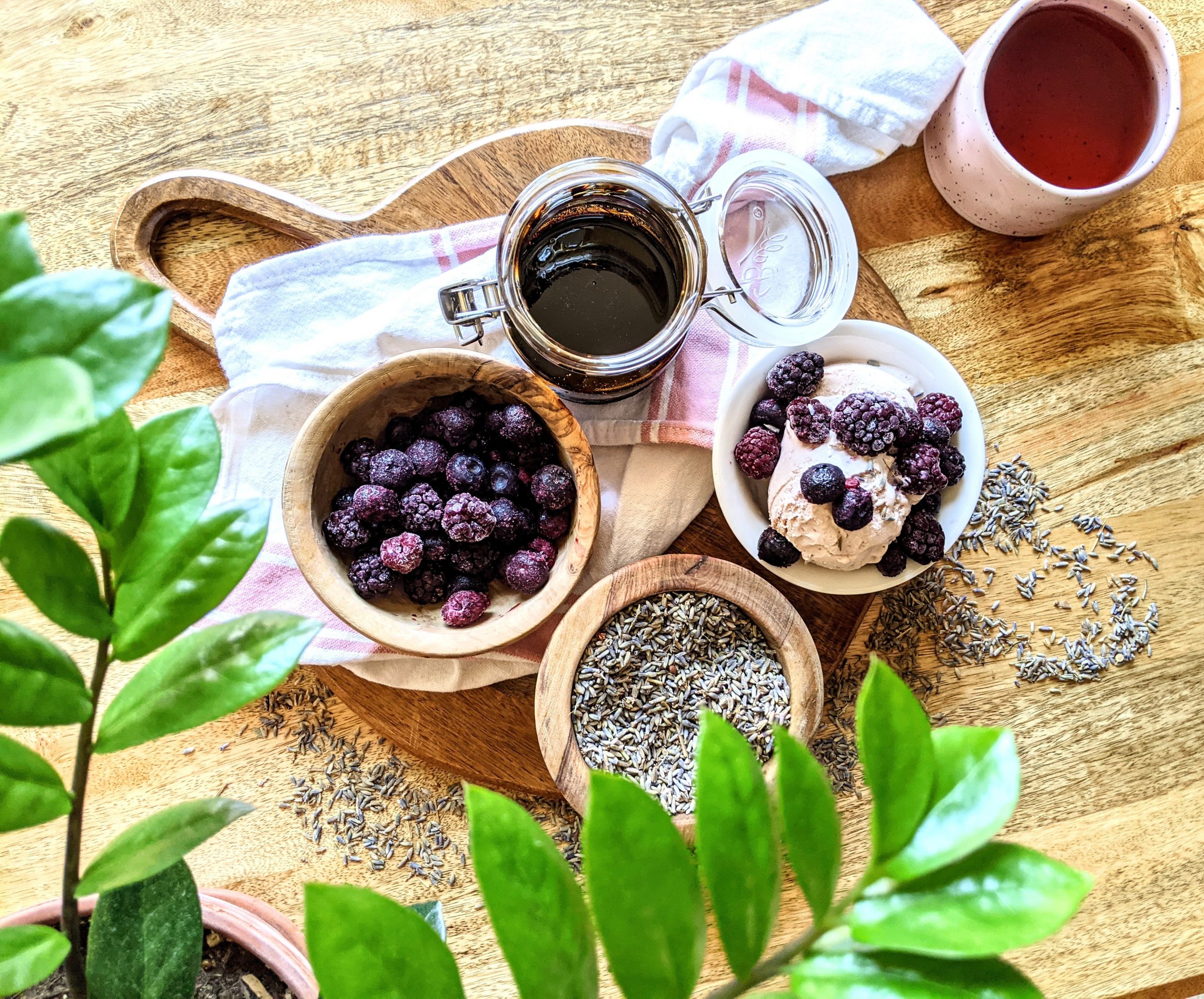 A sundae bar complete with thick balsamic glaze, frozen mixed berries, French lavender, and ice cream. A cup of tea sits on the side and a plant is seen in the frame.
