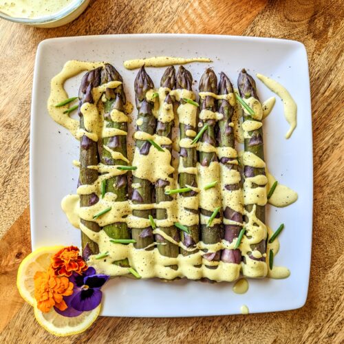 A plate of steamed purple asparagus with tarragon and chive Hollandaise drizzled overtop; garnished with lemon slices and purple and orange edible flowers..