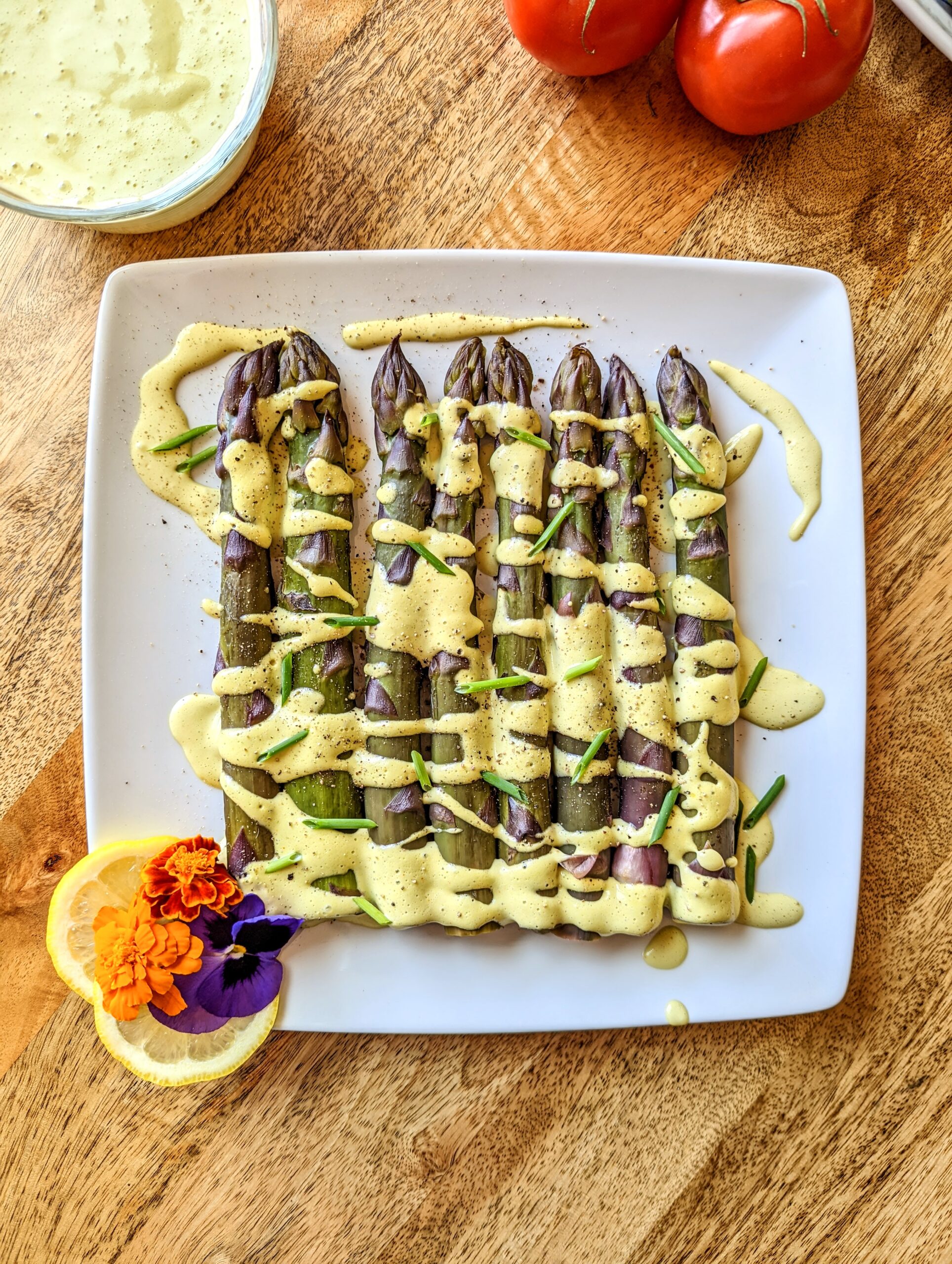 A plate of steamed purple asparagus with tarragon and chive Hollandaise drizzled overtop; garnished with lemon slices and purple and orange edible flowers..