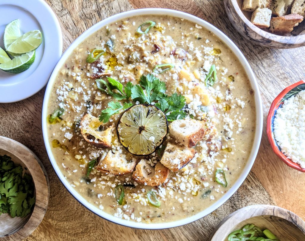 A bowl of Roasted Poblano and Corn Chowder, garnished with homemade croutons, crumbled cotija cheese, cilantro, scallions, and charred lime.