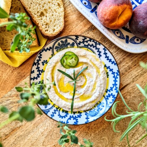 A quirky blue and white plate full of Rosemary and Jalapeño Hummus. The plate is garnished with 3 slices of jalapeño, rosemary needles, and vibrant extra-virgin olive oil. The hummus is frames with fresh bread, peaches, fresh rosemary, and fresh oregano.