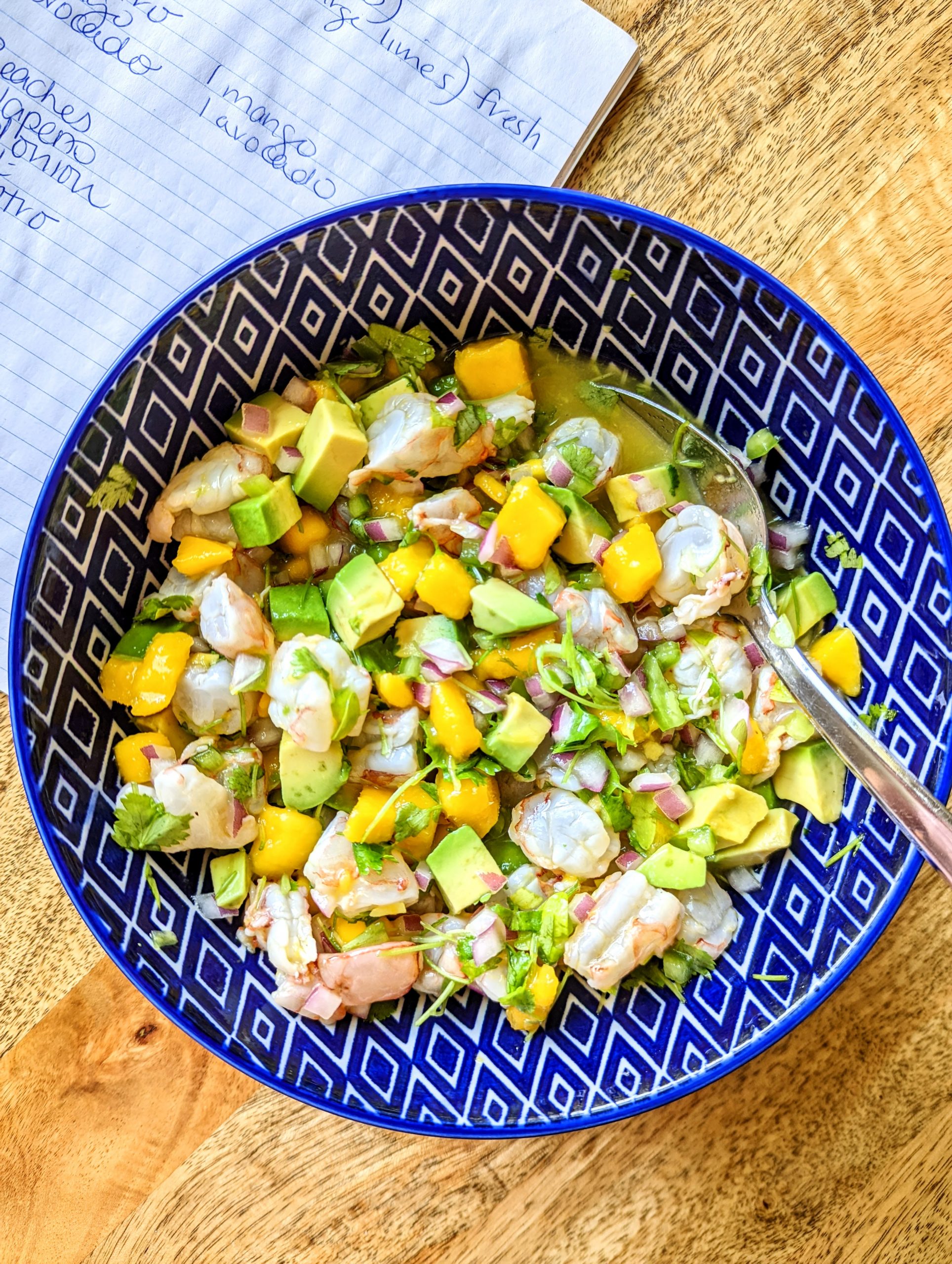 Shrimp ceviche with mango and avocado being mixed together in a bright blue and white bowl. The bowl rests on top of a notebook with part of the recipe written in pen.