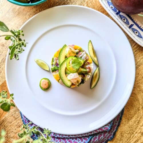 A aerial shot of circular shaped shrimp ceviche with mango and avocado. Two pieces of avocado flank the right side of the plate, and a lime wedge and jalapeno filled with an orange sauce flank the left. A purple and blue napkin cradles the bottom of the plate. Fresh oregano, juices limes, and a mango are visible in the corners of the photo.