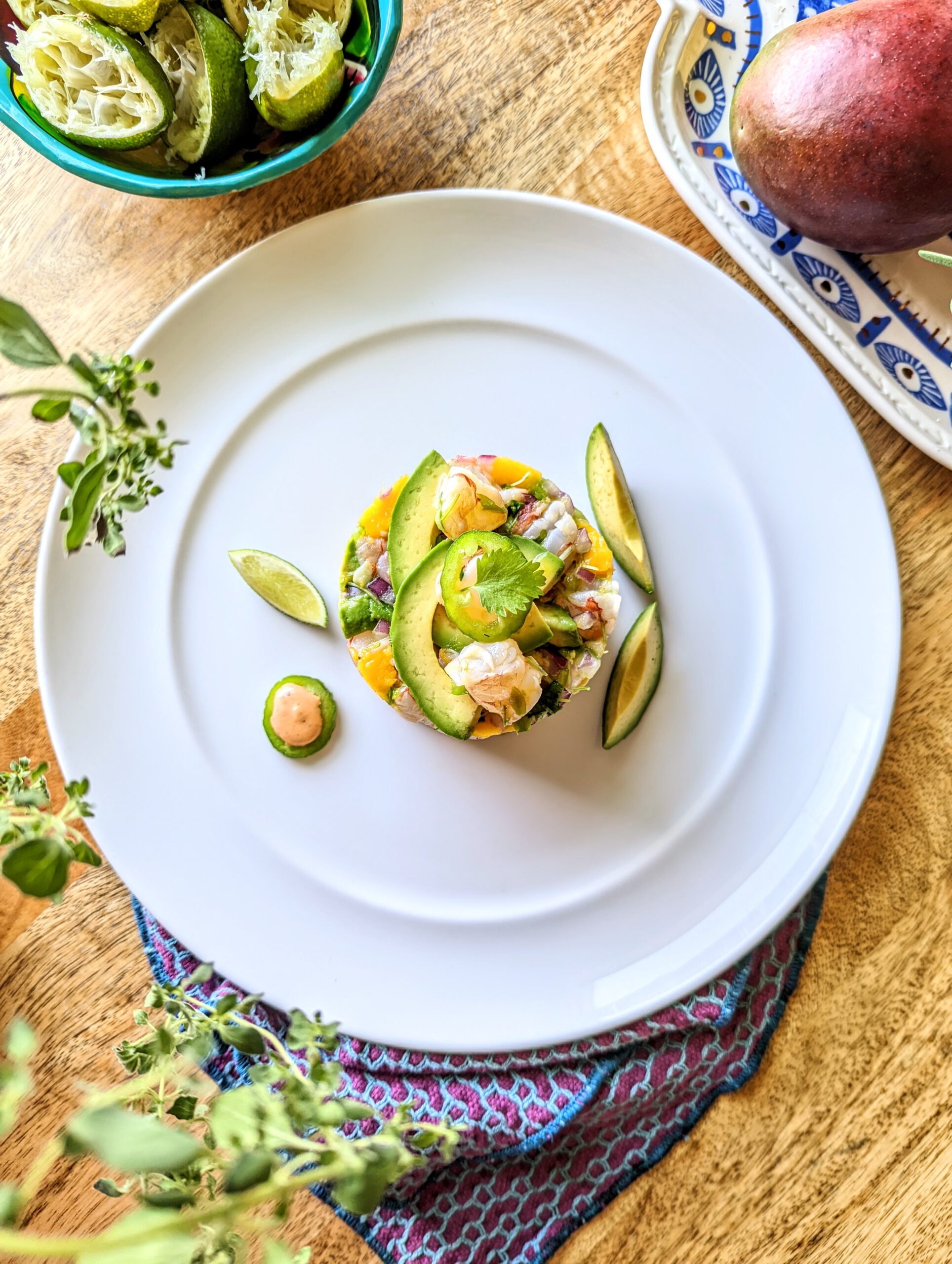 A aerial shot of circular shaped shrimp ceviche with mango and avocado. Two pieces of avocado flank the right side of the plate, and a lime wedge and jalapeno filled with an orange sauce flank the left. A purple and blue napkin cradles the bottom of the plate. Fresh oregano, juices limes, and a mango are visible in the corners of the photo.