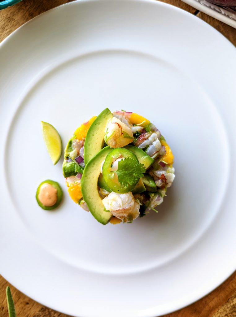 An aerial photo of shrimp ceviche with mango and avocado. Topped with sliced avocado, pieces of shrimp, a jalapeño slice and a stem of fresh cilantro. Serve with lime wedges and spicy sauce.