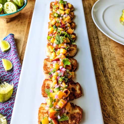 A straight-on view of a white platter of 8 pieces of grilled halloumi cheese topped with peach salsa.