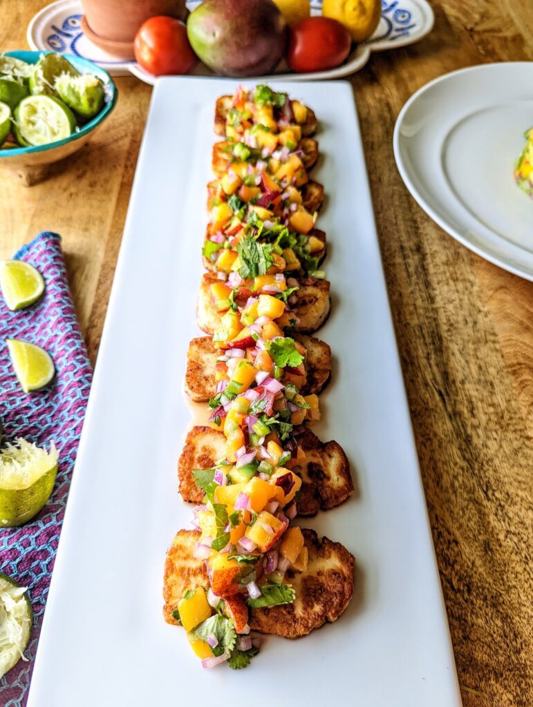 A straight-on view of a white platter of 8 pieces of grilled halloumi cheese topped with peach salsa.