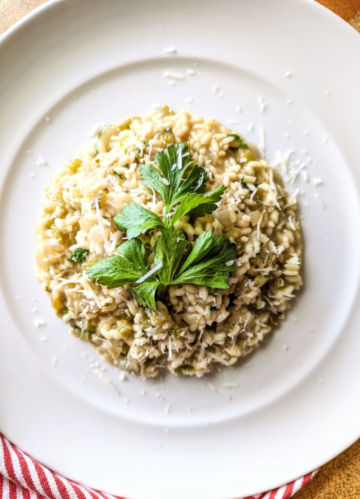 A large white plate of Celery and Tarragon Risotto garnished with celery leaves and Pecorino Romano. You can see a red and white kitchen towel in the bottom left hand corner.