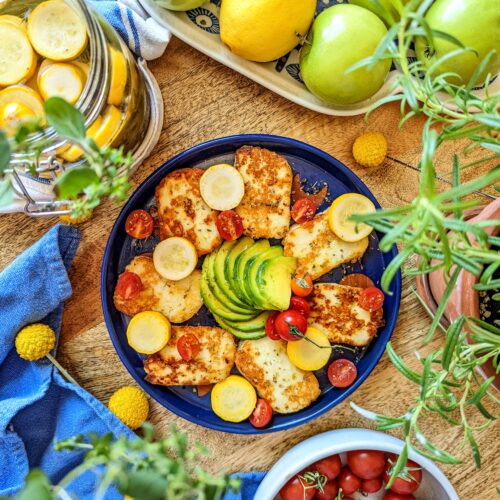 An aerial flat-lay of an indigo-blue plate of grilled halloumi cheese, pickled summer squash, halved cherry tomatoes, and green avocado. Green apples, lemon, a plate of tomatoes, and a jar or pickled summer squash surround the plate.