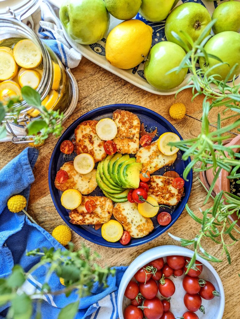 An aerial flat-lay of an indigo-blue plate of grilled halloumi cheese, pickled summer squash, halved cherry tomatoes, and green avocado. Green apples, lemon, a plate of tomatoes, and a jar or pickled summer squash surround the plate.