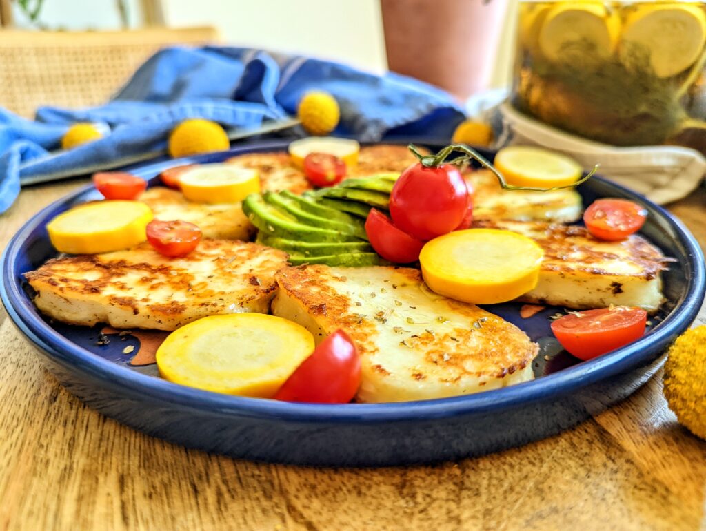 A tableside view or golden brown grilled halloumi cheese, with green avocado, red cherry tomatoes, and yellow pickled summer squash.