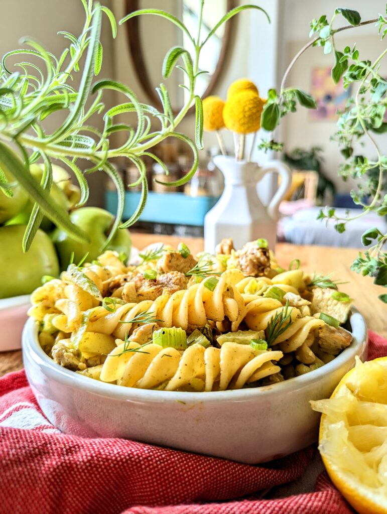 A head-on table view of a shallow bowl of curry chicken and apple pasta salad. Fresh rosemary and oregano plants frame the shot. A bowl of granny smith apples can be seen in the background.