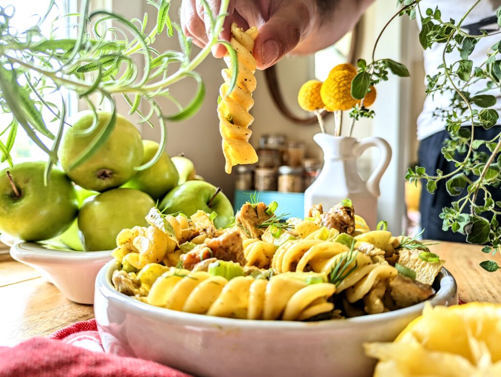 A hand picking up a long fusili noodle of curry chicken and apple pasta salad. The plate is centered in the middle of fresh rosemary and oregano plants. A bowl of granny smith apples can be seen in the background.