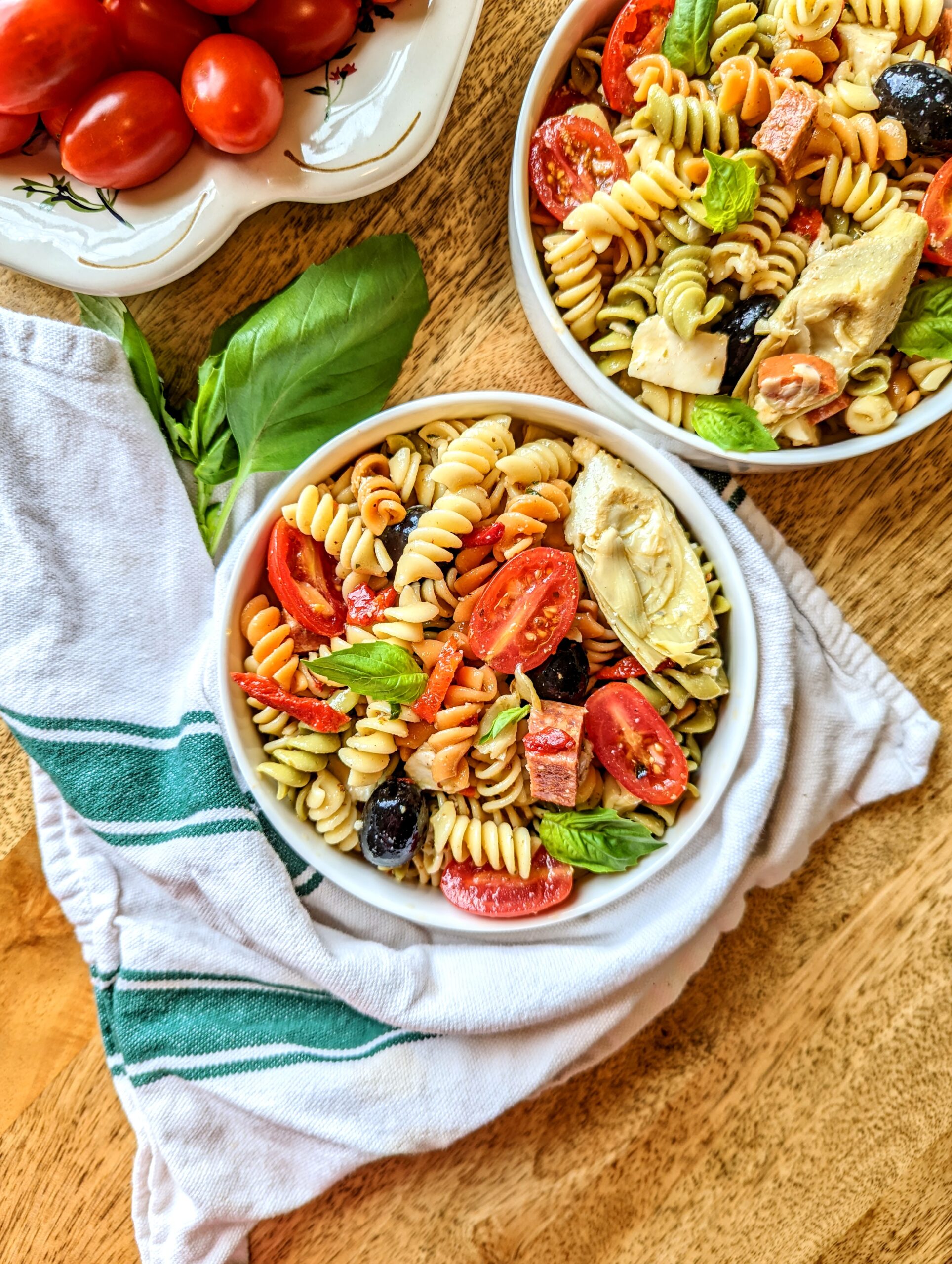 A bowl of loaded antipasto pasta salad, wrapped in a white and forest green kitchen towel.