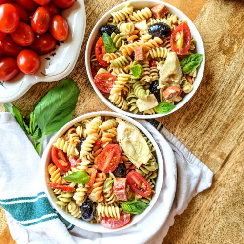 Two bowls of colorful Italian-style antipasto pasta salad. A bowl of cherry tomatoes are in the top left corner. The bottom bowl of pasta salad is wrapped with a green and white kitchen towel.