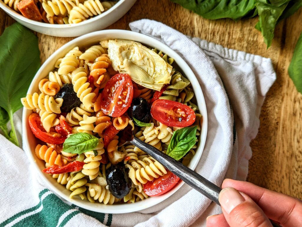 A bowl of loaded antipasto pasta salad with a fork and hand in the bottom right corner.