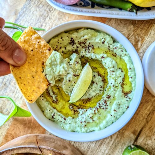 A hand holding a tortilla chip after dipping into the Creamy Avocado Dip with Sumac. The dip is dressed with a generous amount of olive oil and a vibrant green lime wedge.