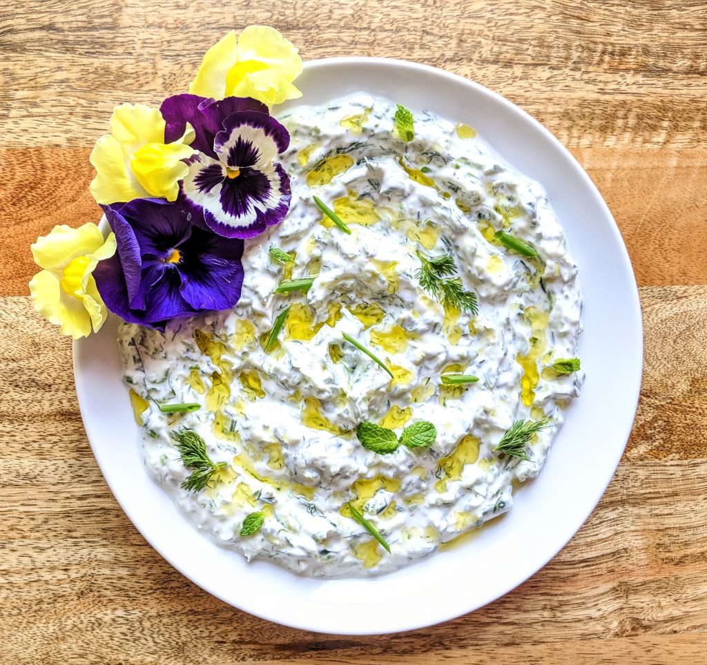 A white plate of yogurt dip with fresh herbs. Purple and yellow edible flowers garnish the top left of the plate. Pops of green from fresh chives, mint, and dill provide contrast.