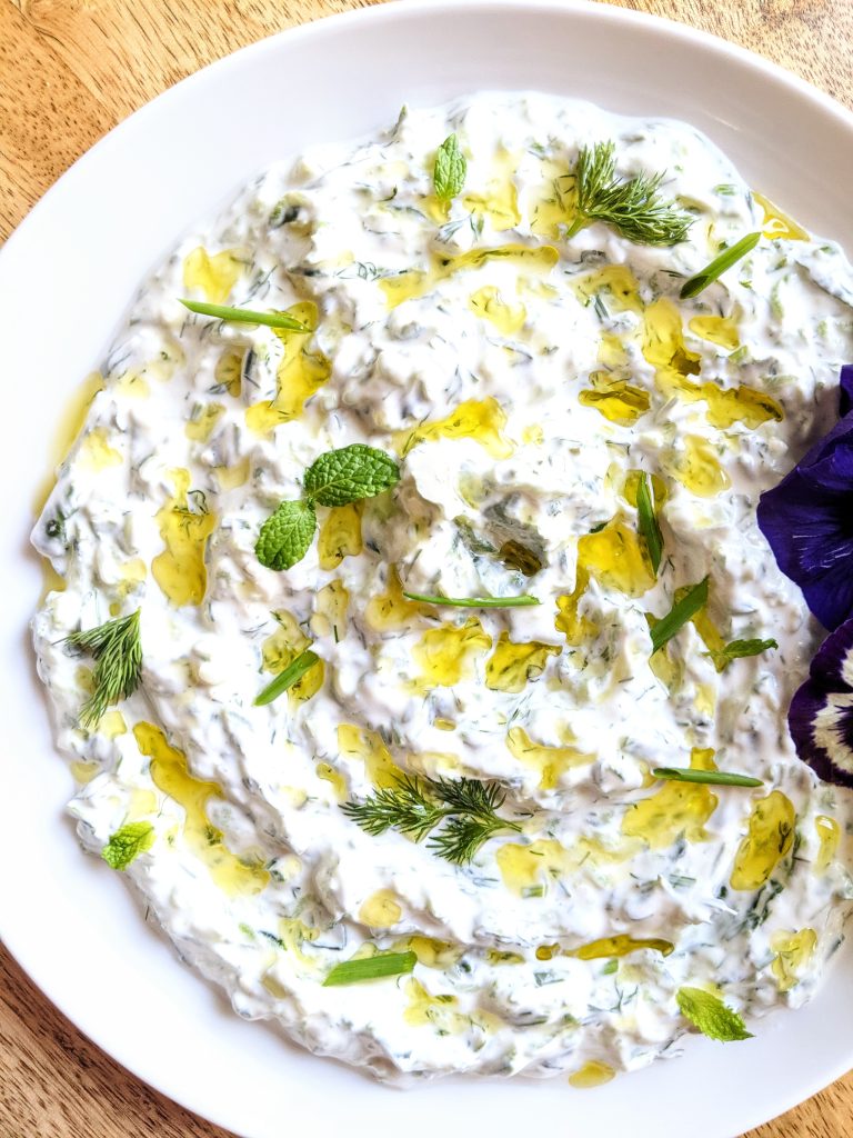 A close-up of a white plate of yogurt dip with fresh herbs. Purple and yellow edible flowers garnish the top left of the plate. Pops of green from fresh chives, mint, and dill provide contrast.