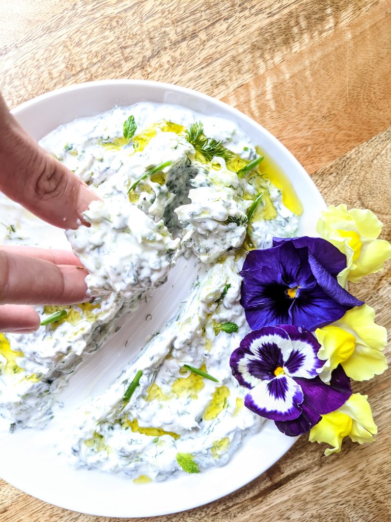 A hand holding a piece of bread, gliding through a plate of Yogurt Dip with Fresh Herbs. Purple and yellow edible flowers can be seen on the right side of the plate.