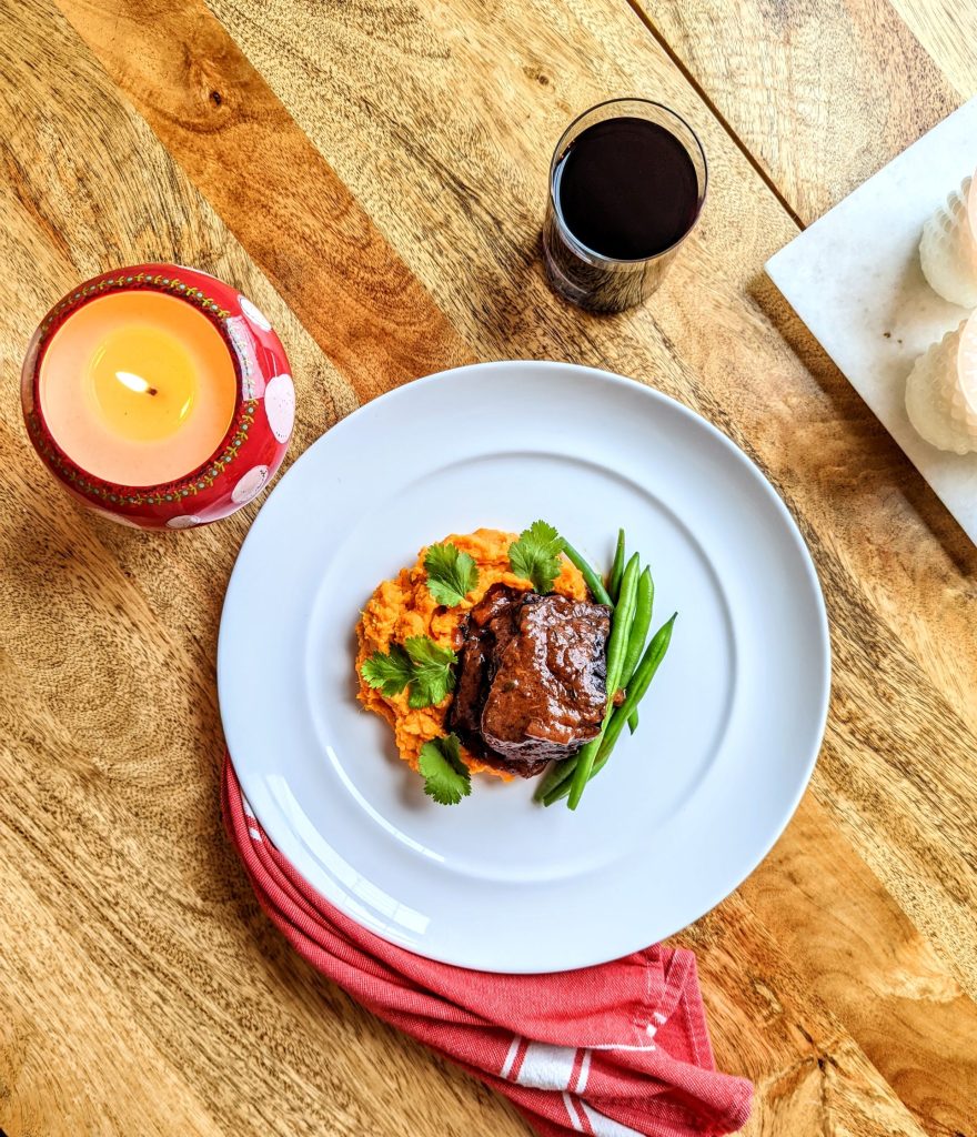 An aerial photo of a white plate of Red Wine Braised Beef Short Ribs. A red and white kitchen towel is draping the bottom of the plate and trailing down the wooden table, a red and white mushroom candle is lit in the top left of the frame, and a short glass of red wine in the top right of the frame.