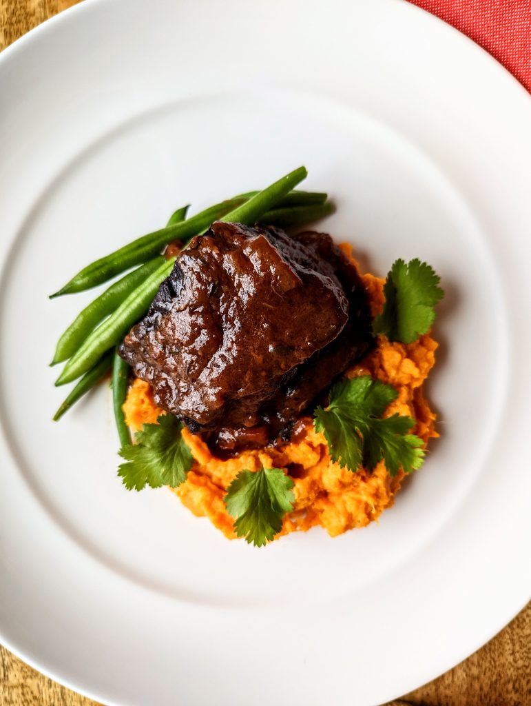 A closeup of Red Wine Beef Short Ribs, served with mashed sweet potatoes, steamed green beans, and garnished with parsley.