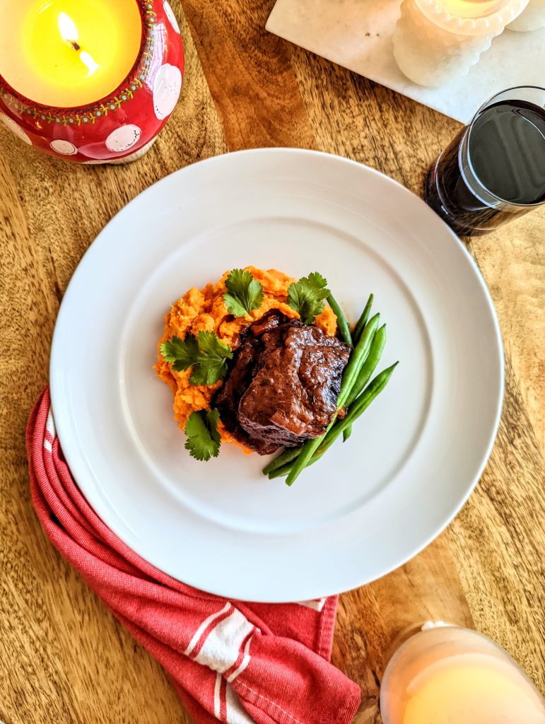 A red and white candle shaped like a mushroom illuminates a plate of Red Wine Braised Beef Short Ribs.