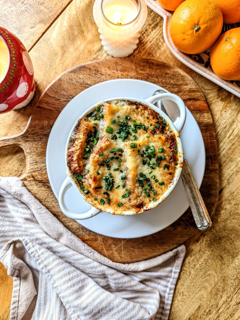 A bowl of French Onion Soup, topped with melted and brown cheese, and fresh chives. A couple of lit candles and a plate of oranges can be seen in the photo.