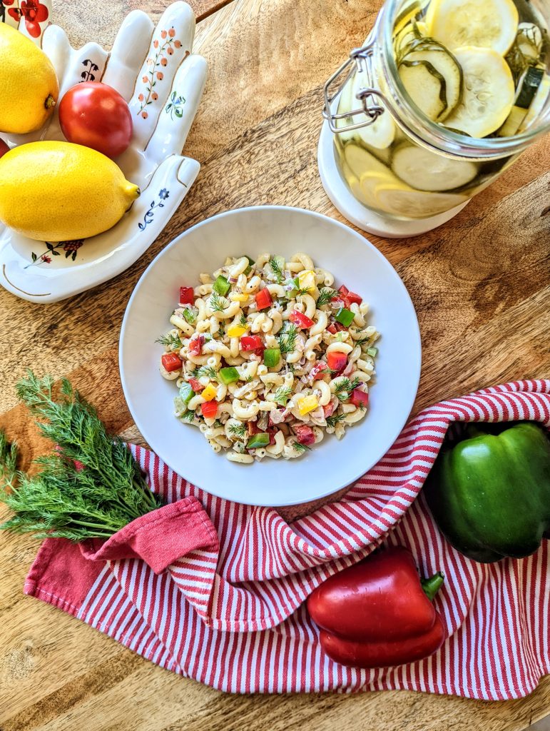 A white bowl of Tuna Pasta Salad with Bell Peppers. Green and red bell peppers, fresh dill, lemon, tomato, and homemade pickles can be seen in the image.