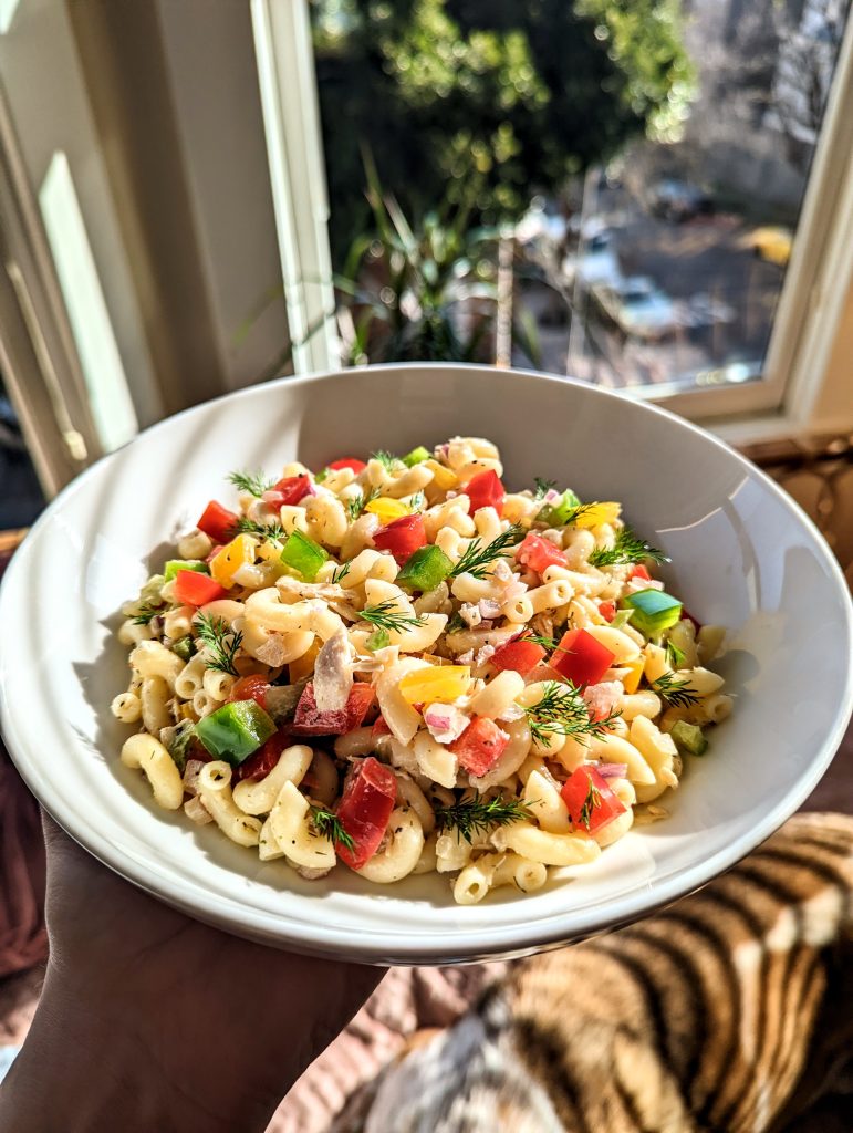 A brightly colored bowl of cold pasta salad with red, green, and yellow bell peppers. The bowl is basking in the bright afternoon light coming through the bay windows.
