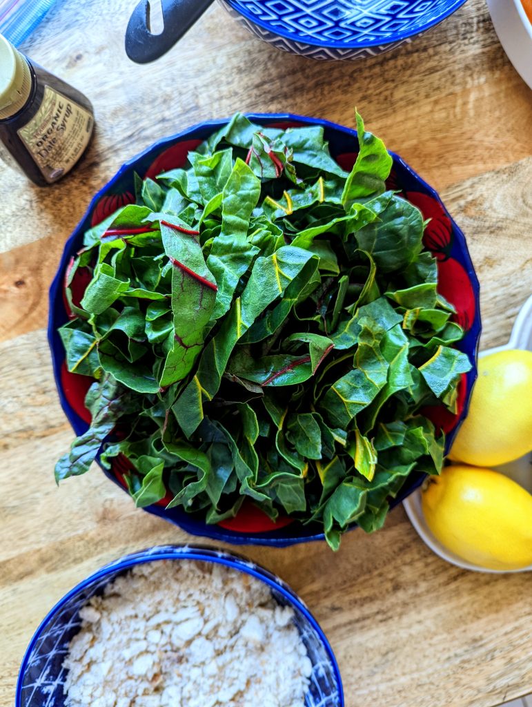 A red and blue bowl of strips of rainbow Swiss chard. Two lemons, a container of date syrup, and breadcrumbs are seen in the photo.