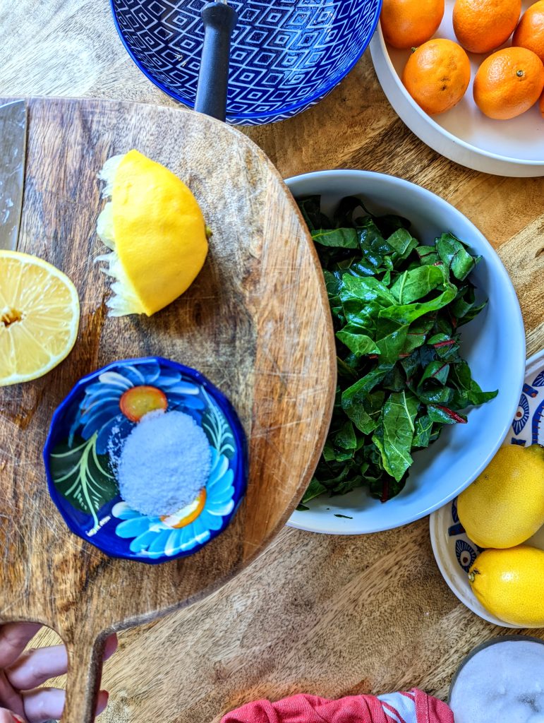 A wooden cutting board with two halves of a lemon and a small blue bowl of salt. A large white bowl of dark, leafy greens can be seen under the cutting board.