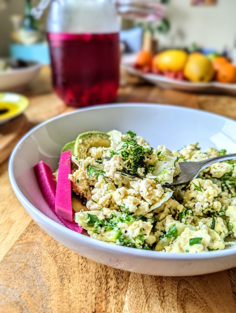 A forkful of Creamy Kale Scramble with Fresh Herbs. Two vibrant pink pickled turnips and sliced avocado can be seen on the plate as well.