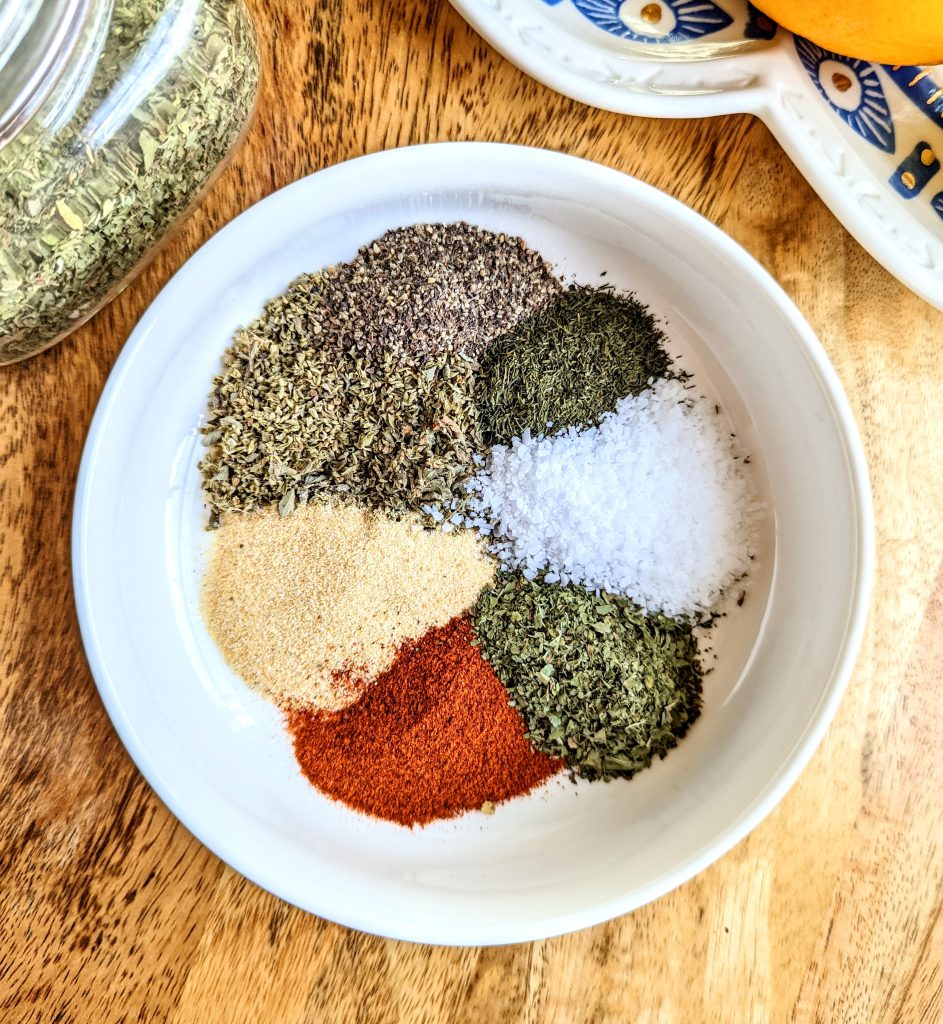 A white bowl full of colorful spices. Smoked paprika, garlic powder, oregano, black pepper, dill, kosher salt, and mint.