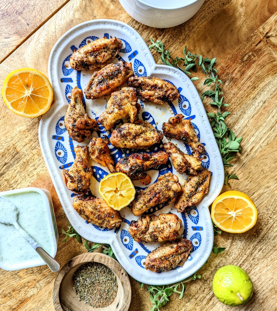 A large blue and white platter of chicken wings. Sprigs of fresh oregano, lemon halves, a small bowl of spice mix, and a bowl of feta and mint dip are visible.